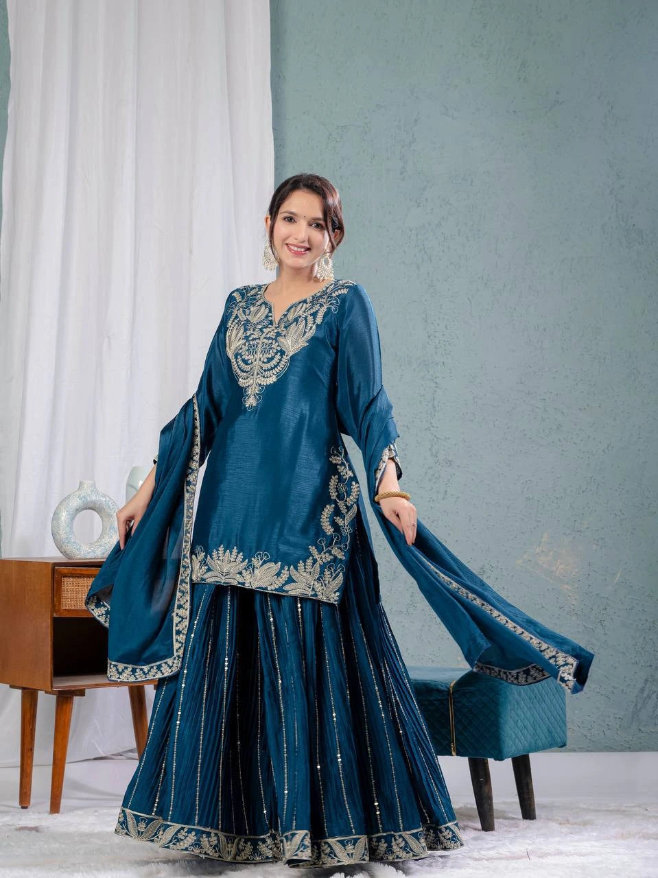Woman in a blue traditional outfit with intricate designs in a room setting.